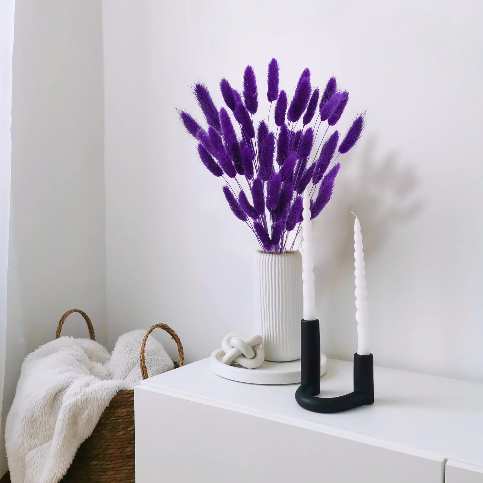 Purple bunny tail grass in a white vase with two white candles in a black holder on a white surface.