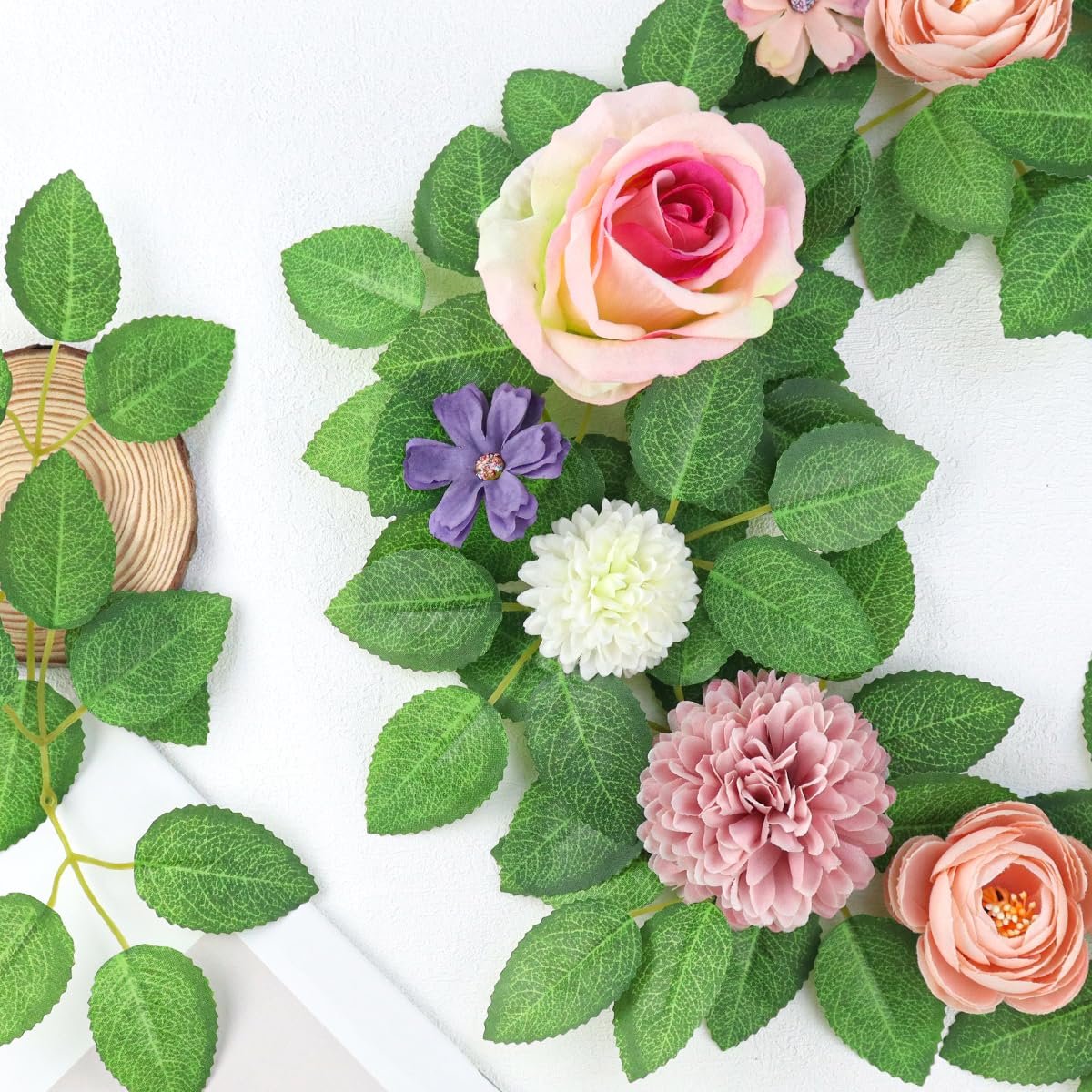 Artificial green rose leaves and assorted silk flowers arranged on a white background with a wood slice.