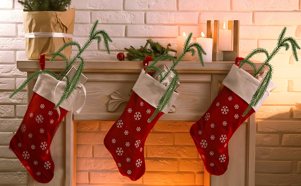 Three red Christmas stockings with snowflake patterns hanging on a mantelpiece, adorned with artificial pine branches and lit candles.