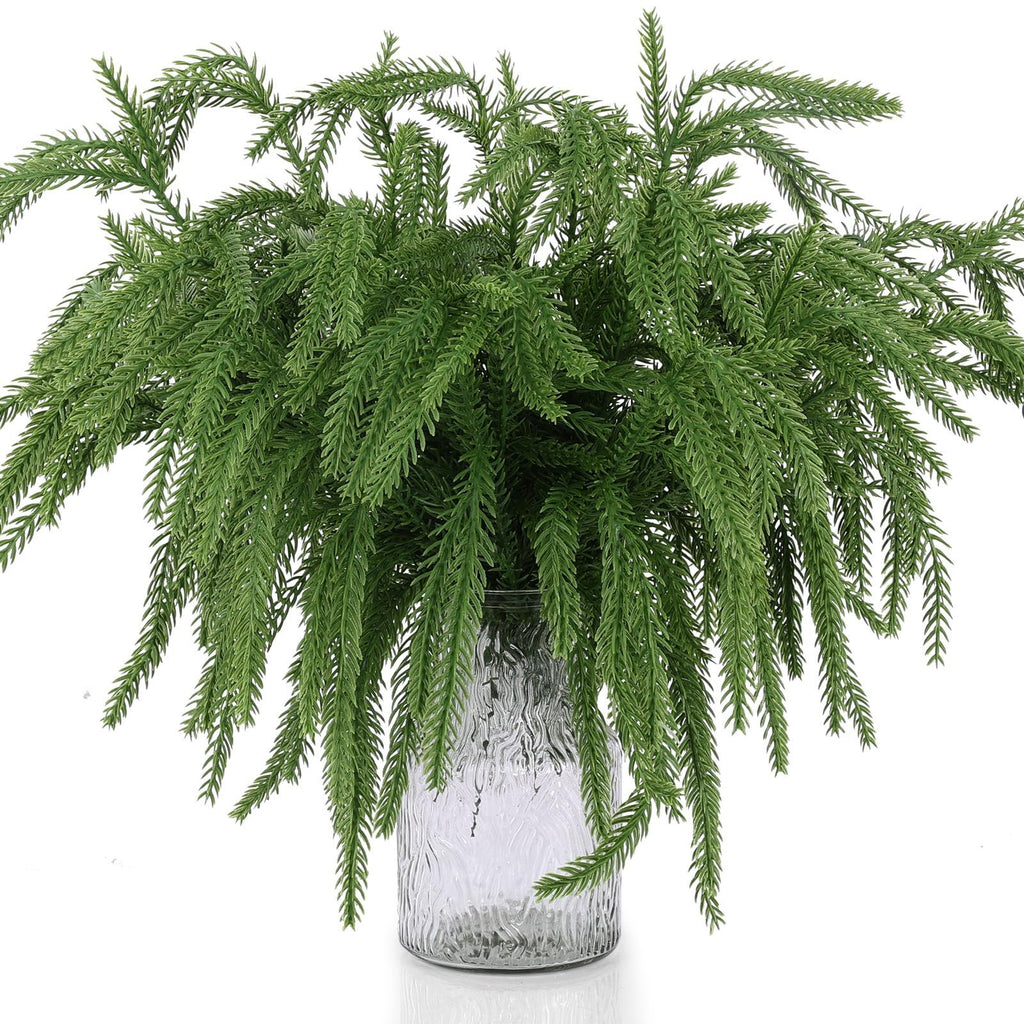 Artificial Norfolk pine branches arranged in a textured glass vase against a white background.