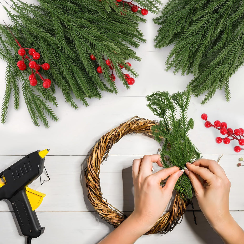 Hands crafting a wreath with artificial pine branches and red berries, using a hot glue gun on a white wooden surface.