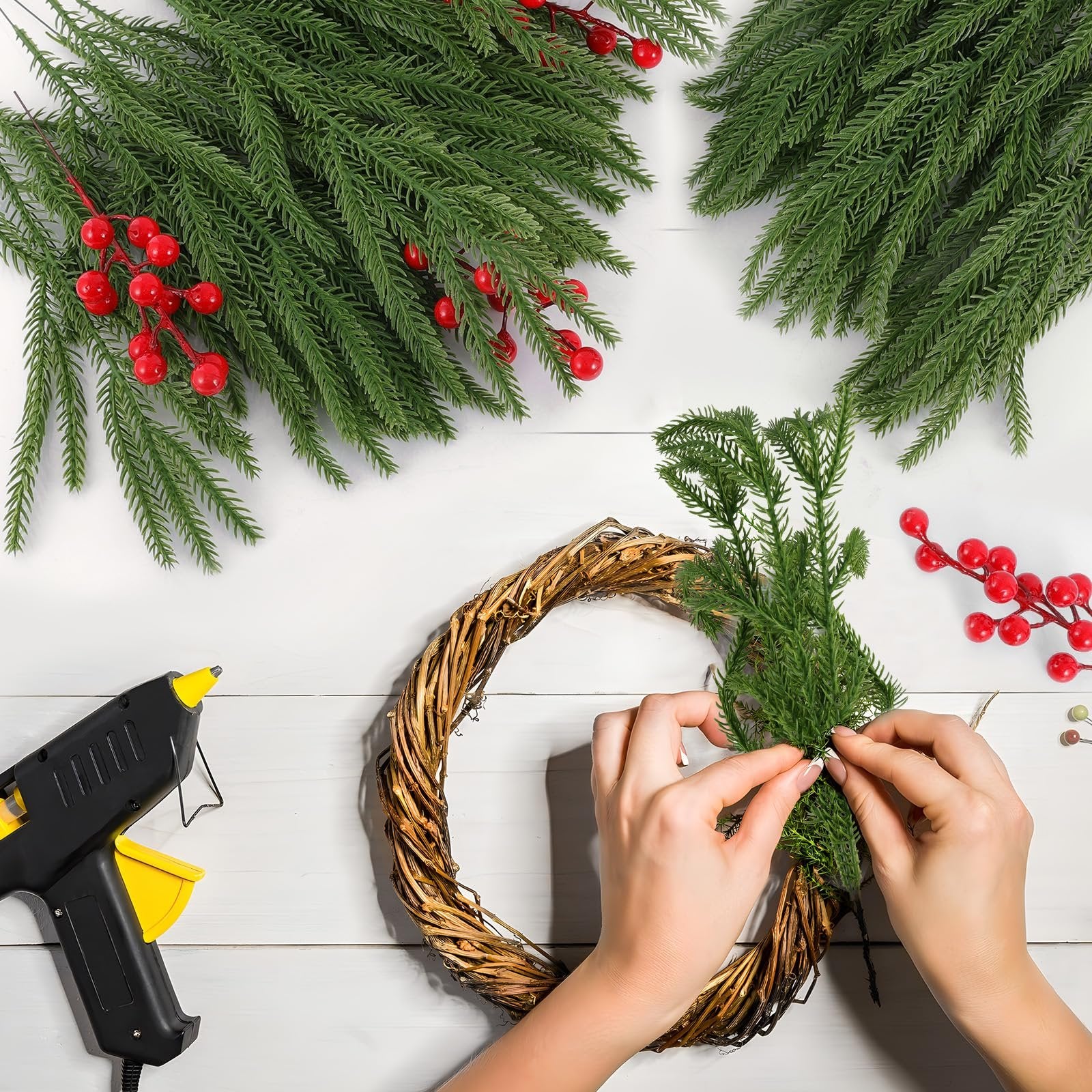 Hands crafting a wreath with artificial pine branches and red berries, using a hot glue gun on a white wooden surface.