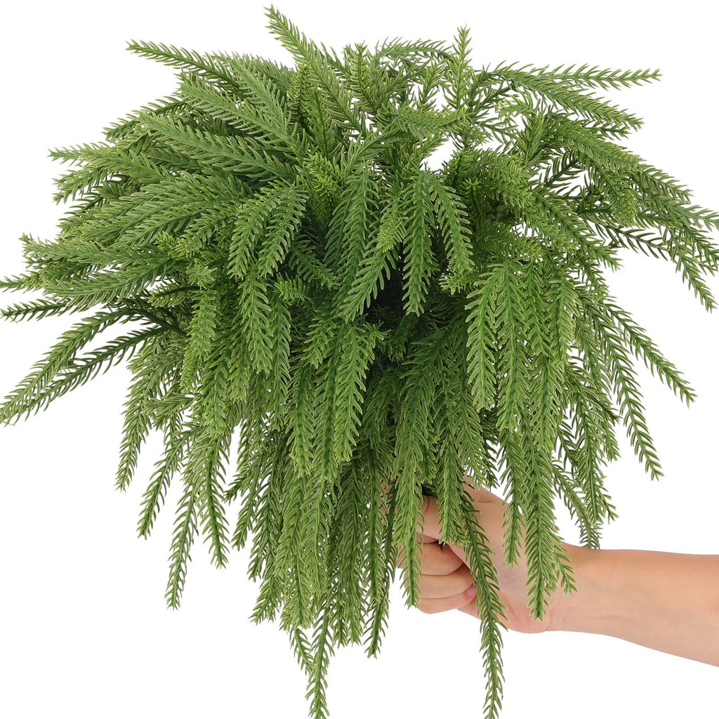 Hand holding a bunch of artificial green Norfolk pine picks with detailed needles against a white background.