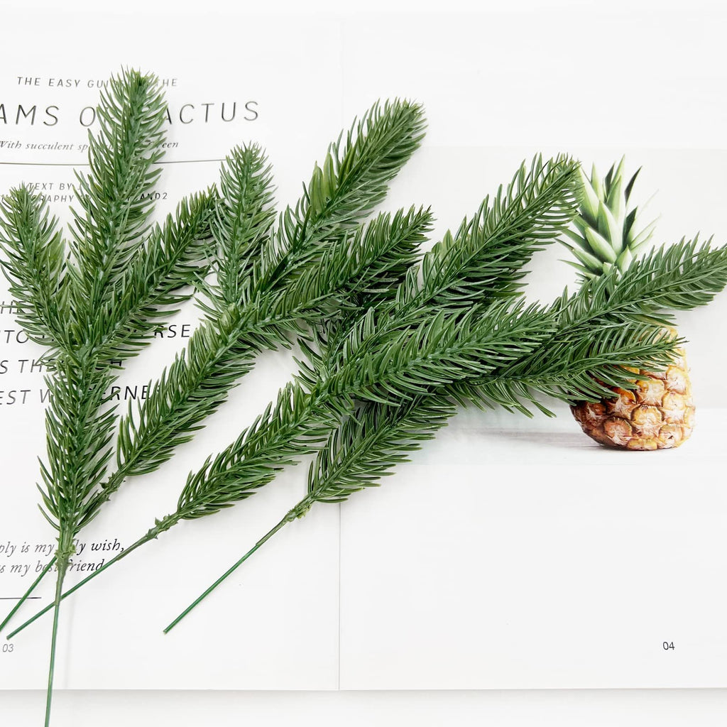 Artificial pine branches arranged on a white surface with a pineapple in the background.