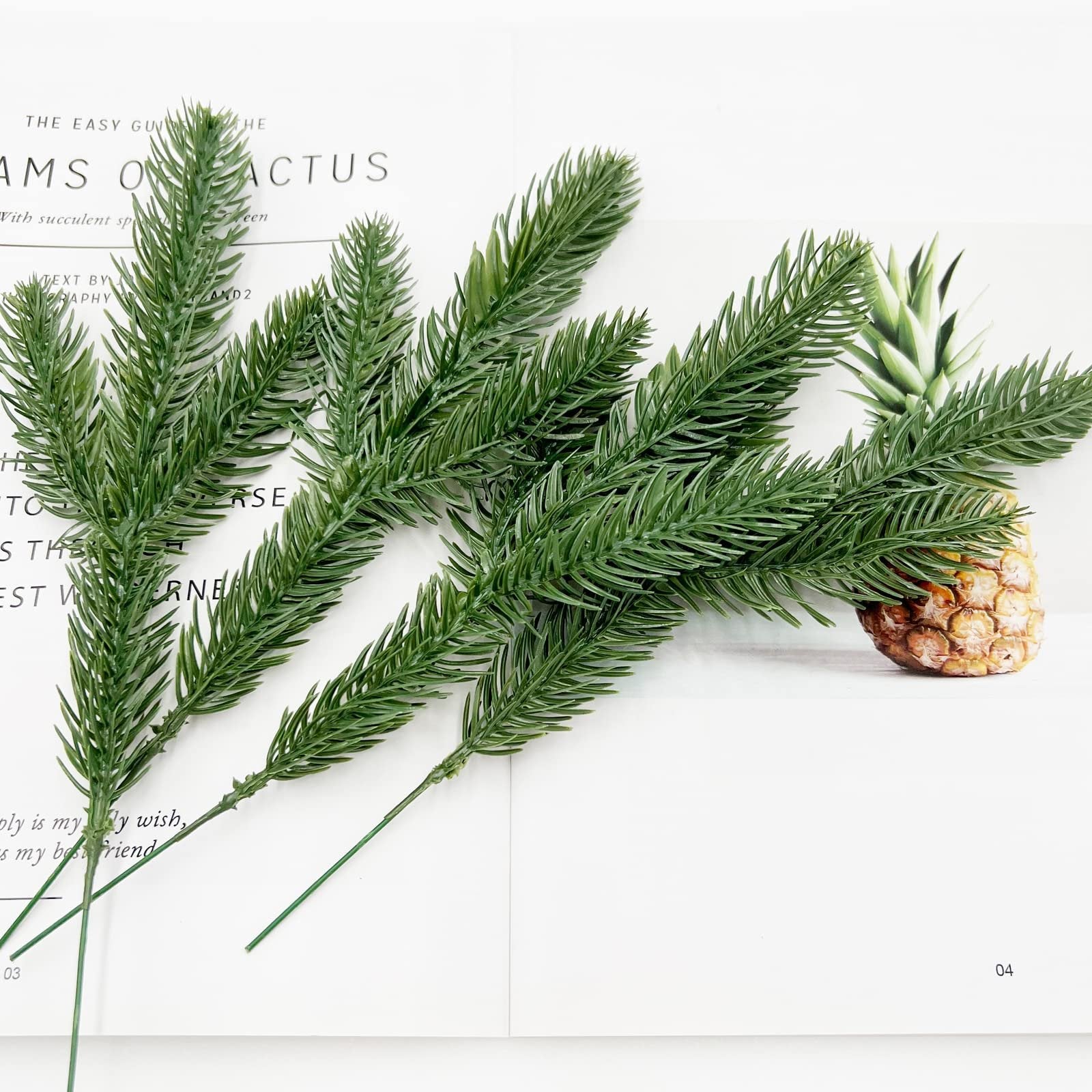 Artificial pine branches arranged on a white surface with a pineapple in the background.