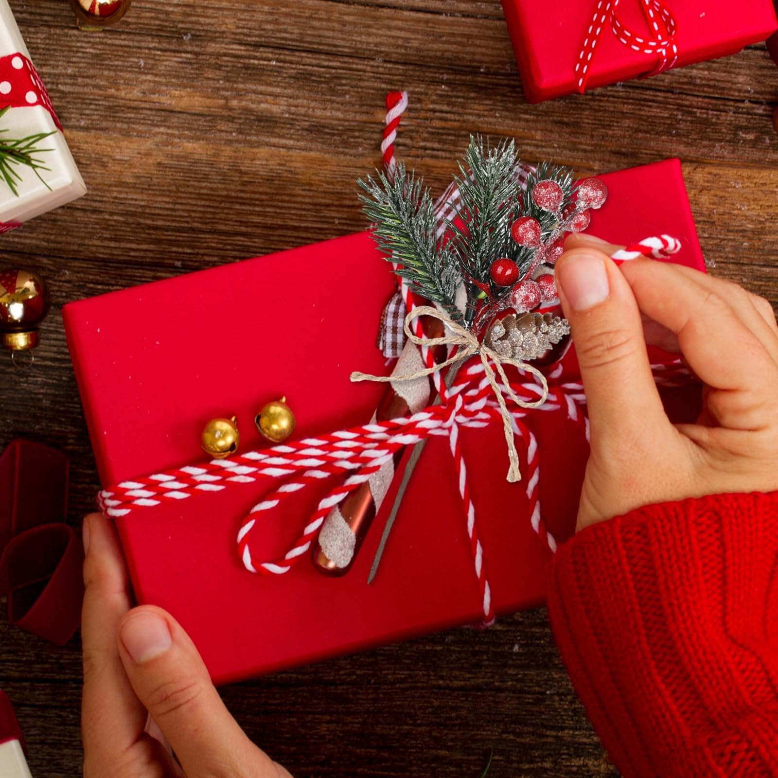 Hands decorating a red gift box with faux cedar, pine needles, berries, and pinecones for Christmas.