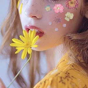 Woman with pressed flowers on her face holding a yellow daisy near her mouth