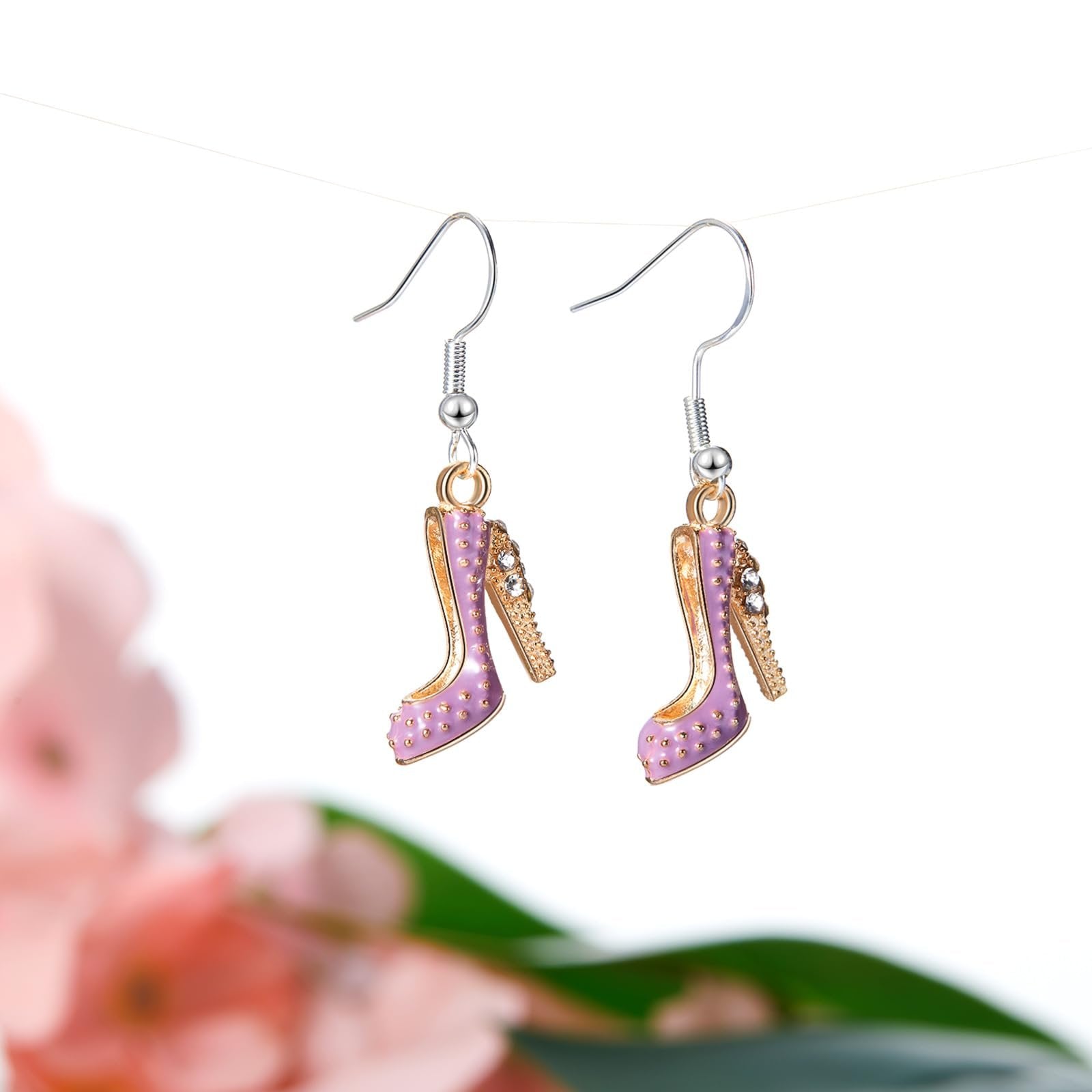 Pair of purple high heel charm earrings with gold accents and rhinestones, hanging from a wire against a blurred floral background.