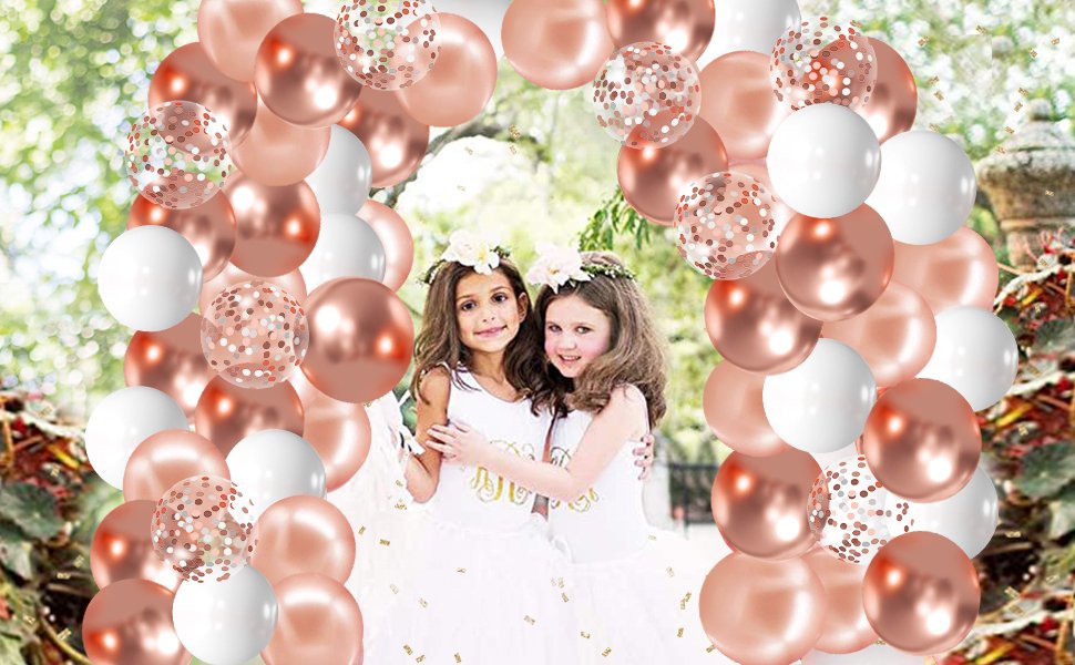 Two young girls in white dresses with flower crowns, embracing under a rose gold balloon arch with confetti and white balloons.