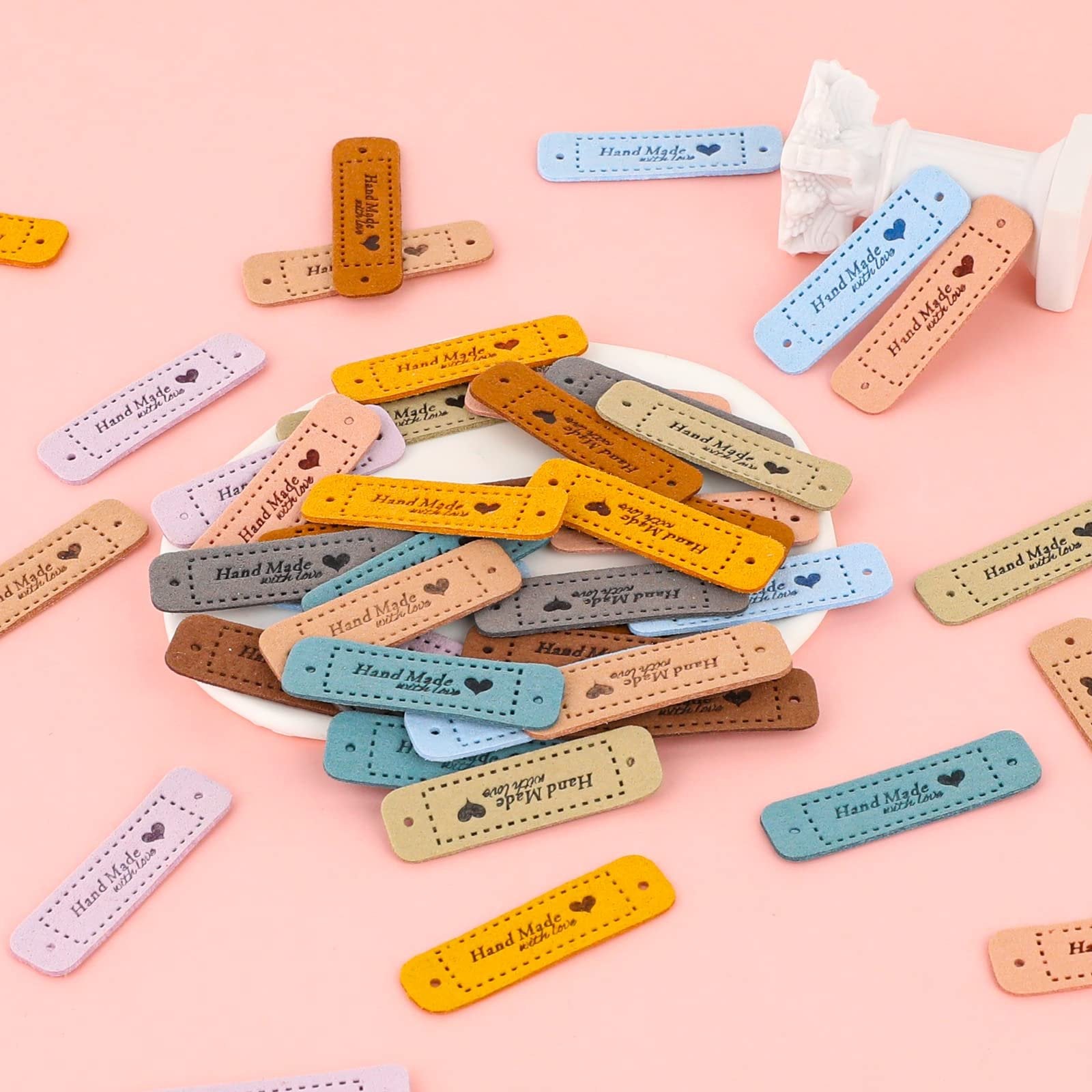 Assortment of 10-color faux leather handmade tags with holes for crochet projects, scattered on a pink surface.