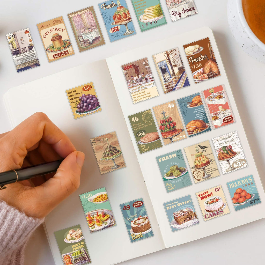 A hand holding a pen over a notebook filled with vintage food postage stamp stickers. The stickers feature various food items like cakes, grapes, and roasted chicken.