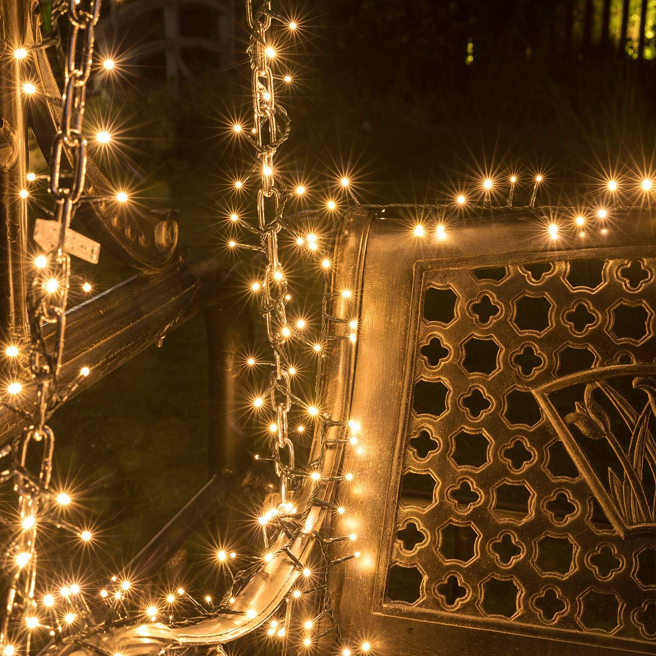 Cluster string lights with warm white LEDs draped over a metal chain and ornate bench in the dark.