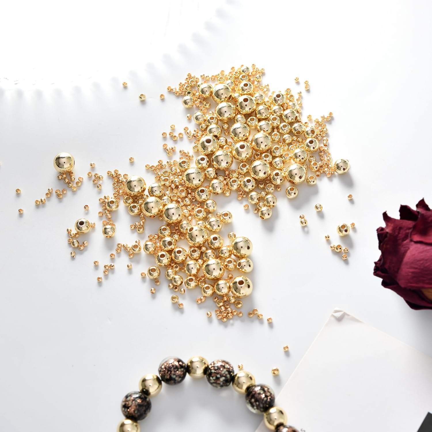 Assortment of gold-filled brass round spacer beads scattered on a white surface with a dried rose and bracelet.