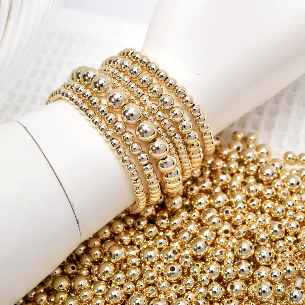 Gold-filled round spacer beads in various sizes, some strung into bracelets on a white mannequin arm, others scattered on a textured white surface.