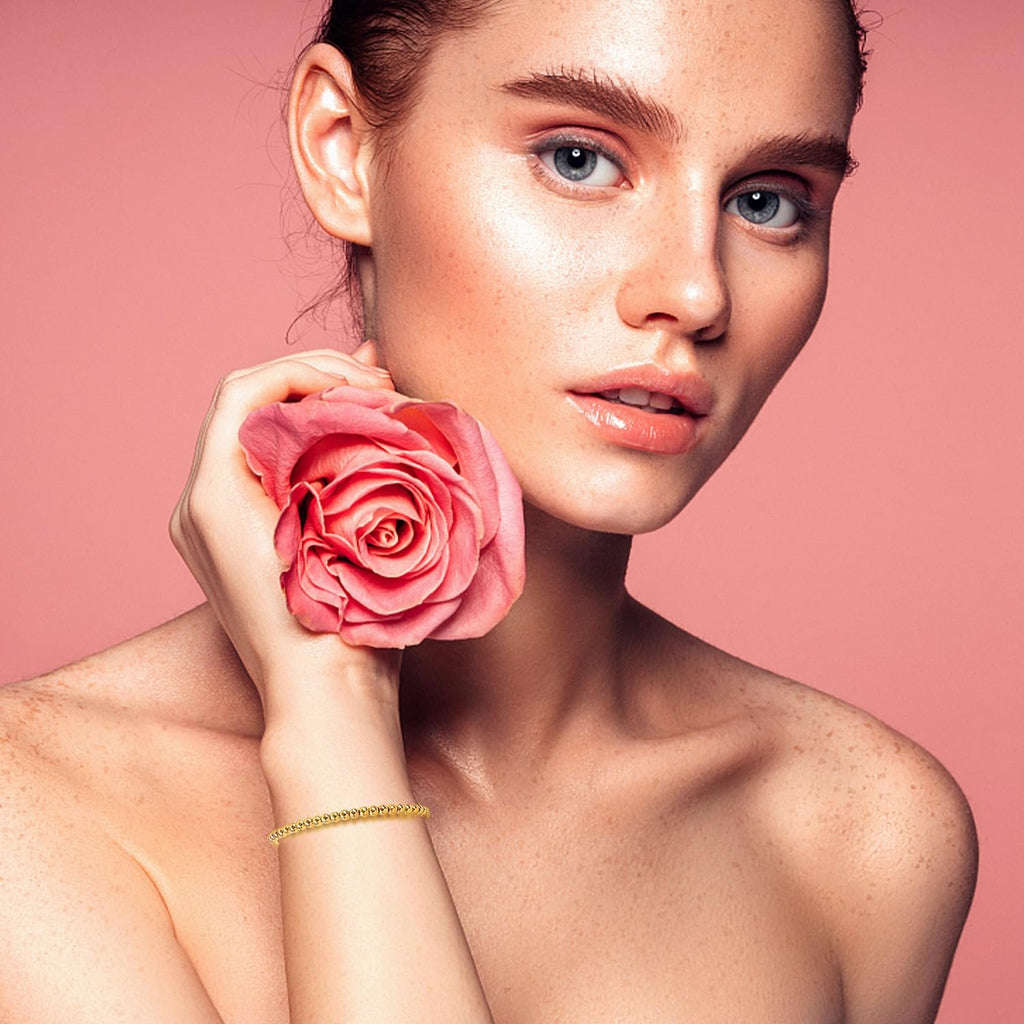 Woman with a pink rose and a gold beaded bracelet on her wrist against a pink background
