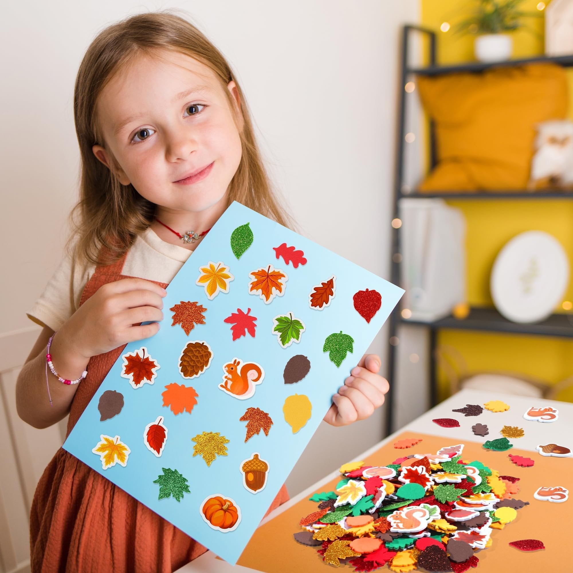 Young girl holding a blue sheet with assorted fall leaf foam stickers, a pile of stickers on a table.