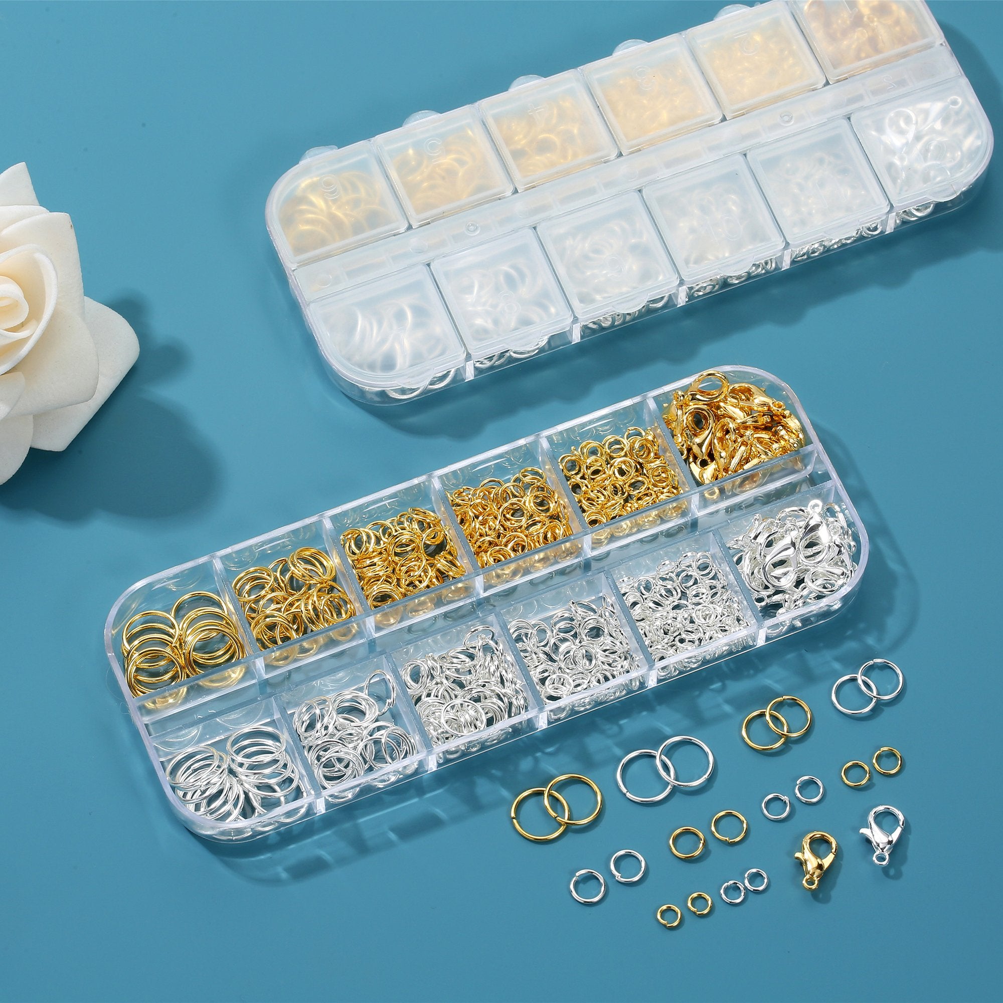 Two clear storage boxes filled with gold and silver jump rings and lobster clasps, with loose rings and clasps scattered on a blue surface.
