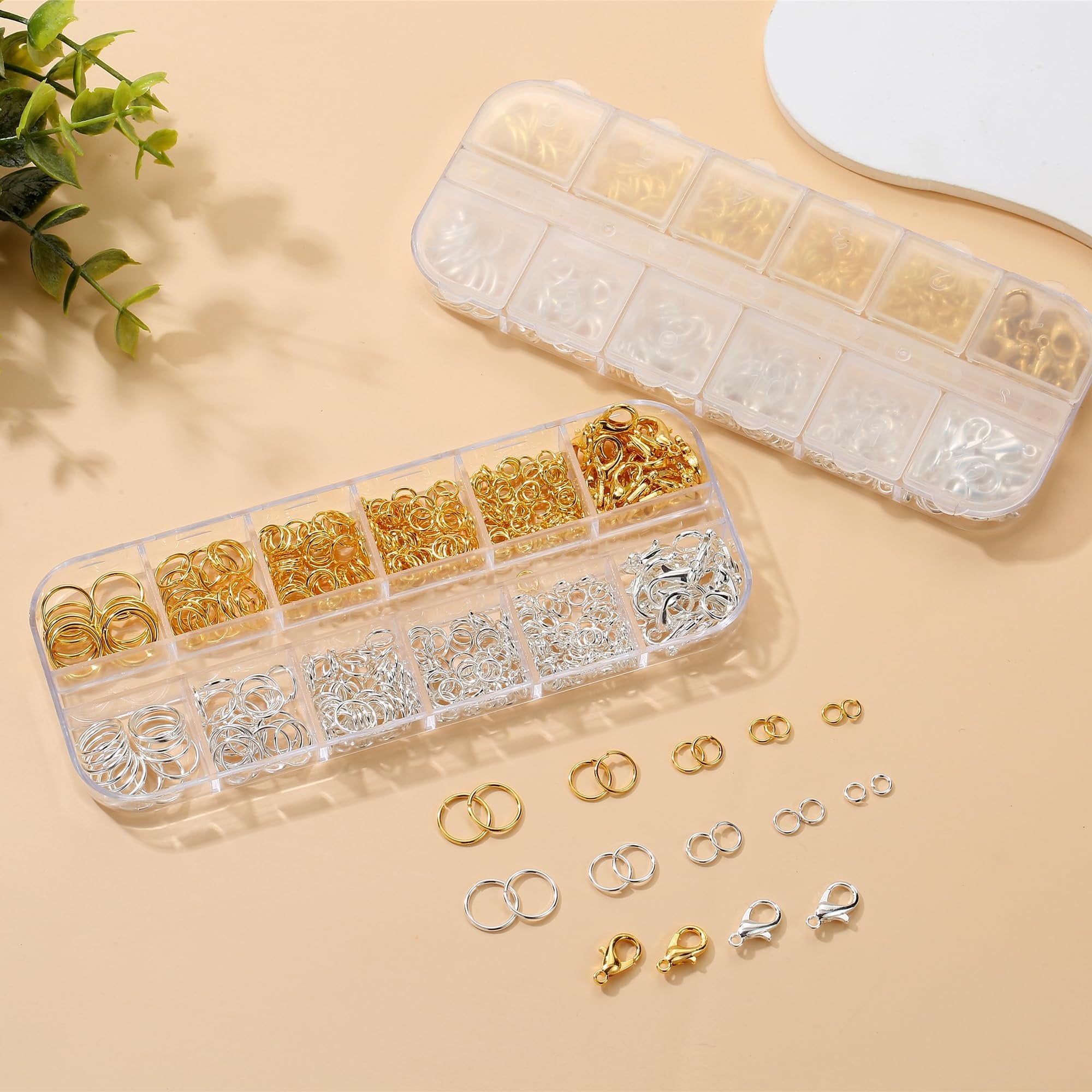 Two clear plastic organizer boxes filled with gold and silver jump rings and lobster clasps, with some rings and clasps scattered on the surface.