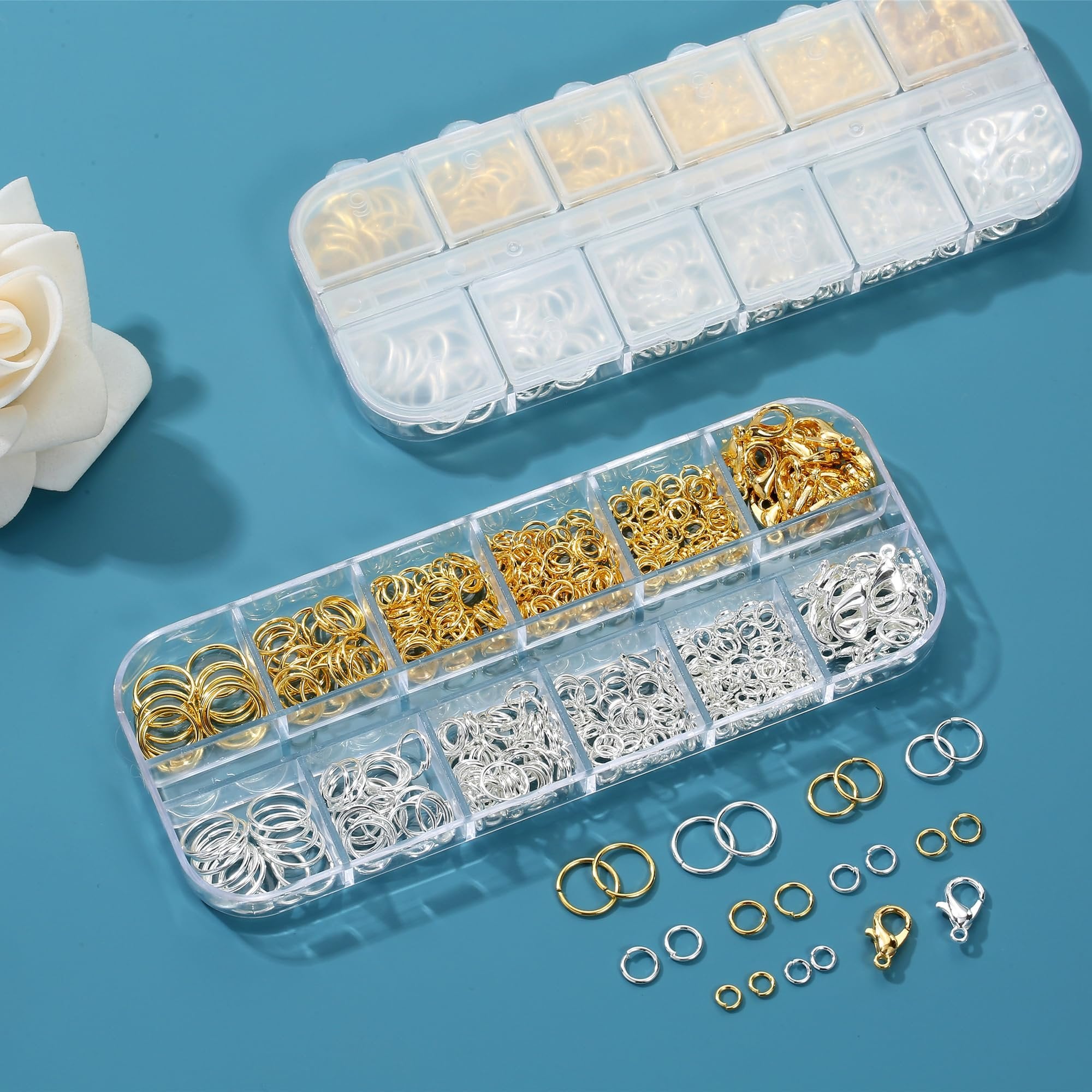 Two clear plastic organizer boxes filled with gold and silver jump rings and lobster clasps, with loose rings and clasps scattered on a blue surface.