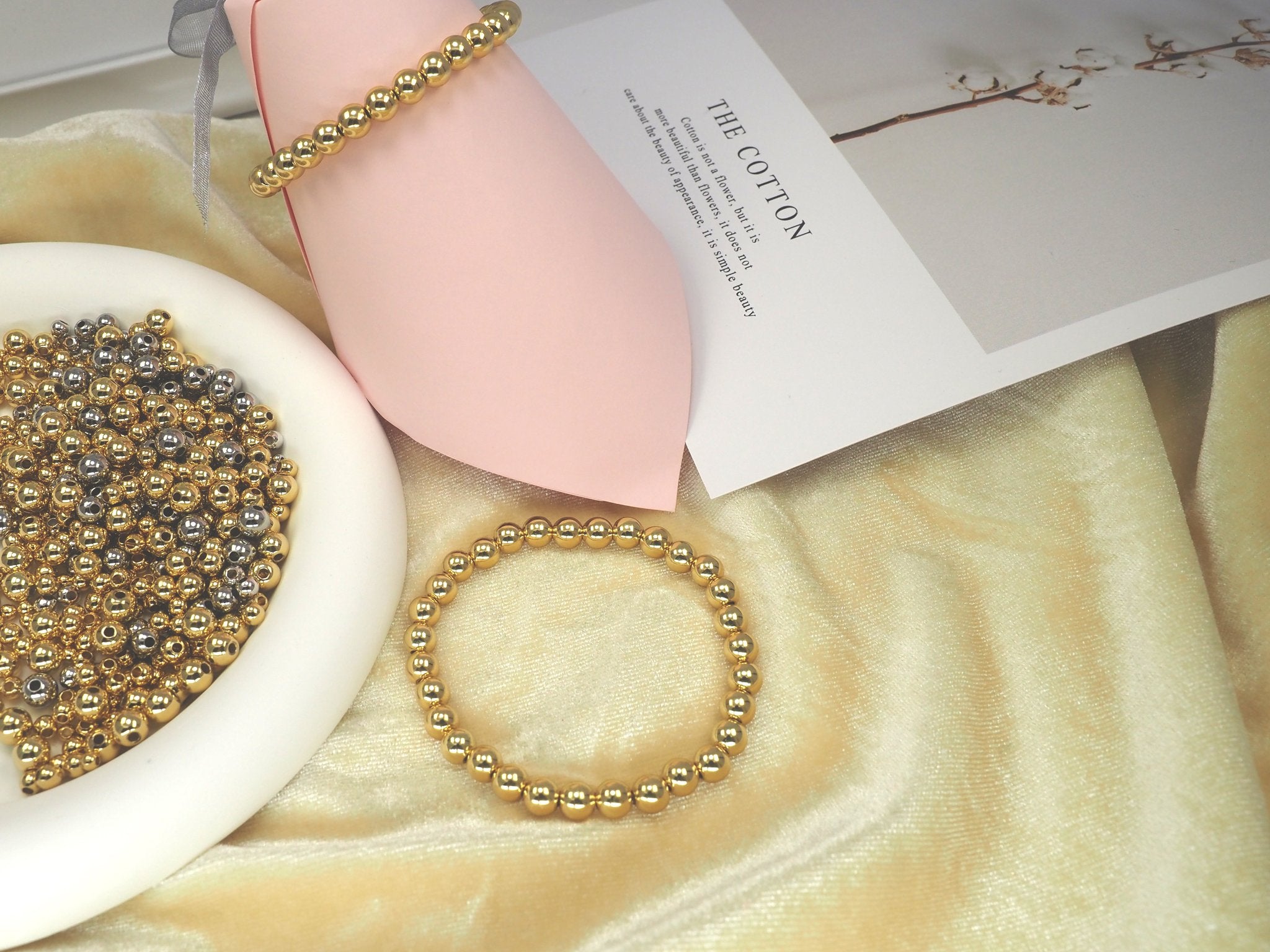 Gold and silver round spacer beads in a white bowl and on bracelets, next to a card with text about cotton.