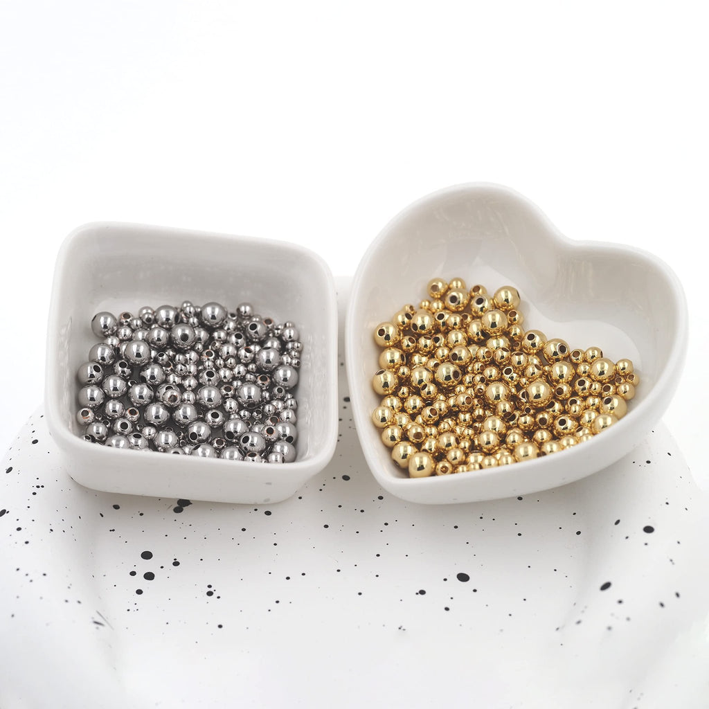 Two white bowls filled with small round spacer beads, one with silver-toned beads and the other with gold-toned beads, on a speckled white surface.
