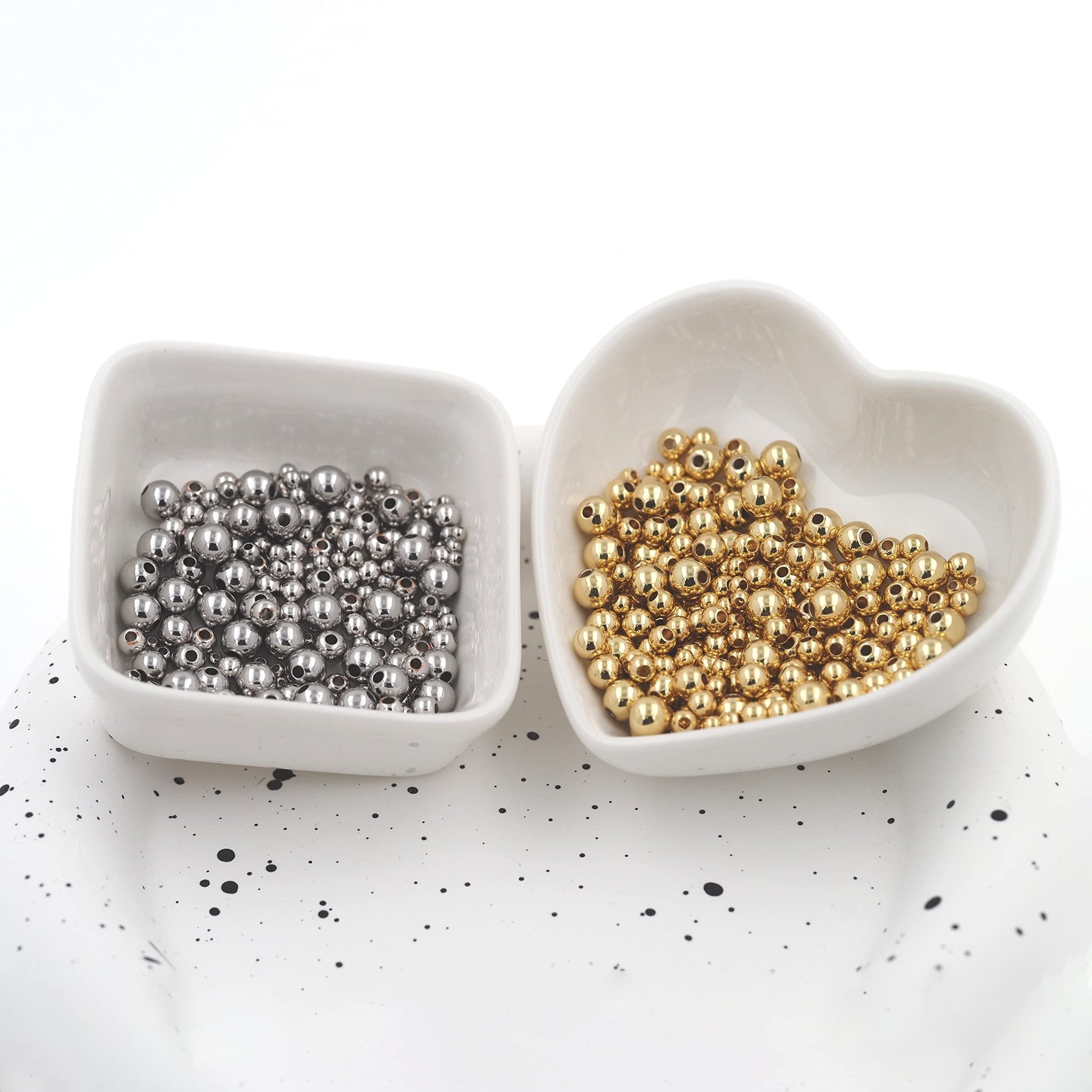 Two white bowls filled with small round spacer beads, one with silver-toned beads and the other with gold-toned beads, on a speckled white surface.