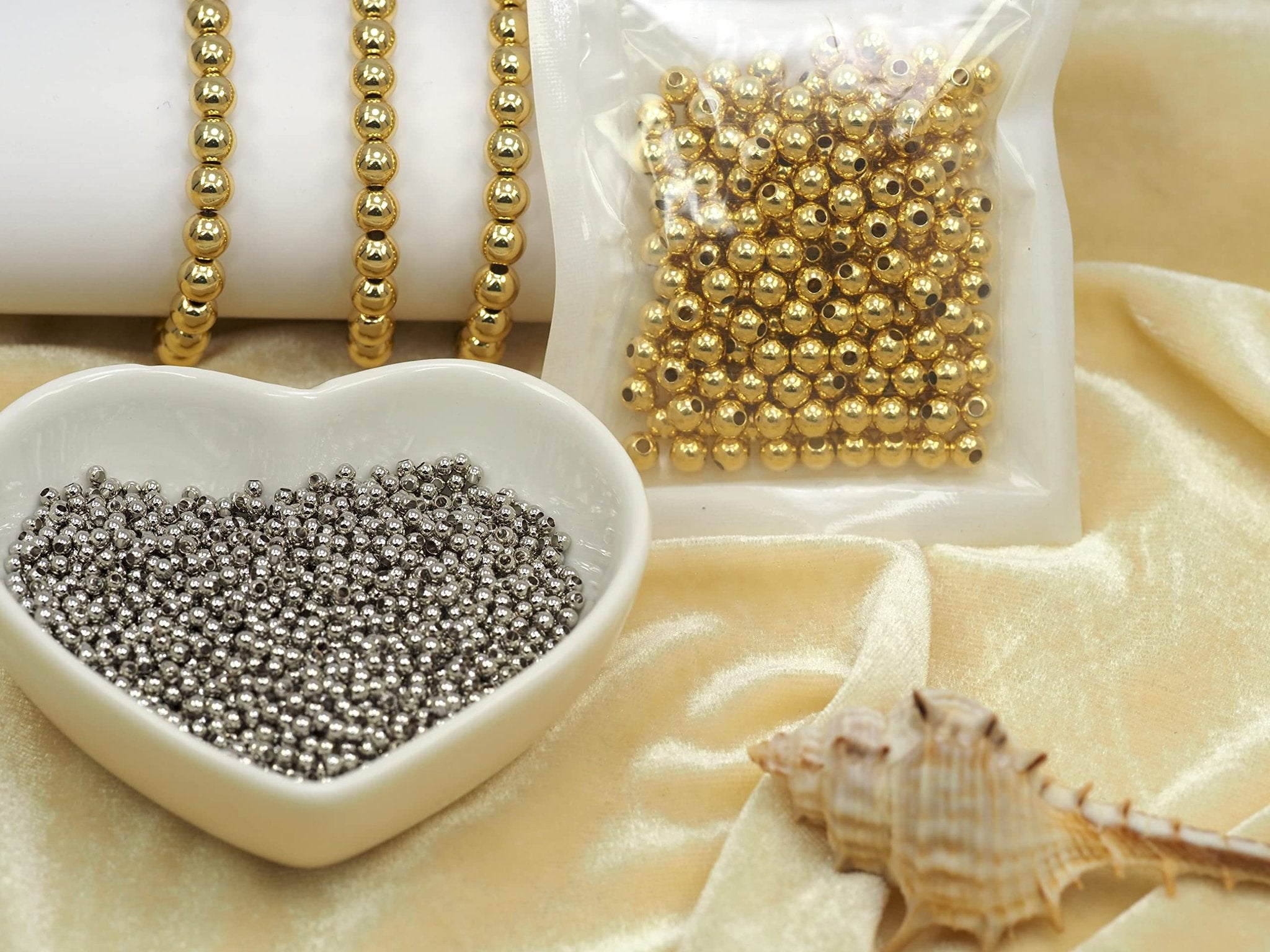 Gold and silver spacer beads in a heart-shaped dish and plastic bag, with gold bead bracelets and a seashell on velvet fabric.