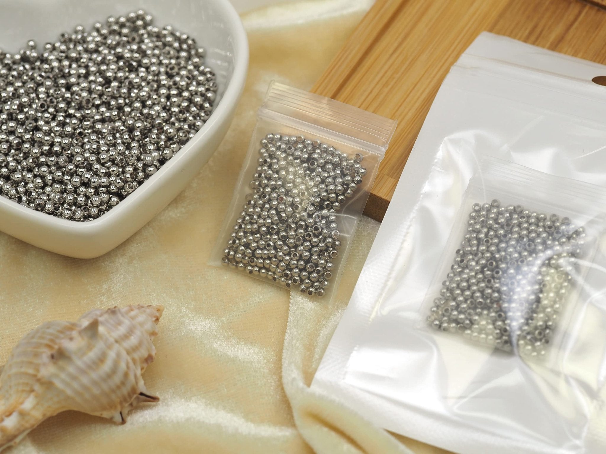 Small silver round spacer beads in a white bowl and two clear plastic bags, with a seashell on a textured fabric surface.