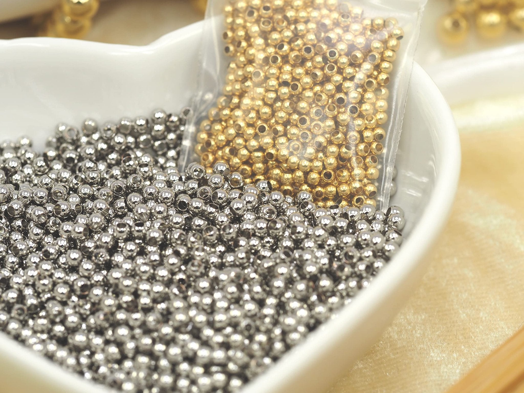 Two bags of small round spacer beads, one silver and one gold, in a white heart-shaped dish.