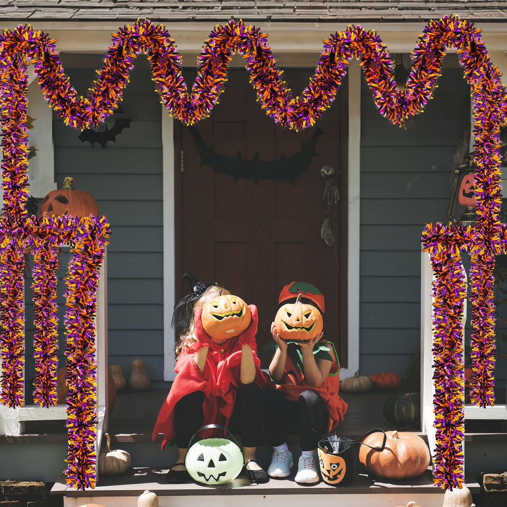 Two children in Halloween costumes sit on porch steps decorated with orange, black, and purple tinsel garland and pumpkins.