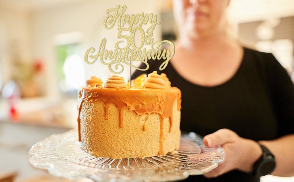 Golden glitter cake topper reading 'Happy 50th Anniversary' on a caramel-frosted cake held by a person.