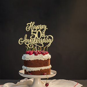 Golden glitter 'Happy 50th Anniversary' cake topper on a two-layer chocolate cake with whipped cream and cherries.