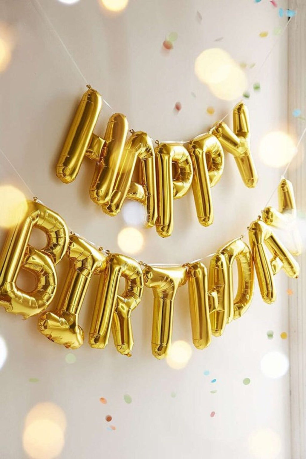 Gold foil letter balloons spelling 'HAPPY BIRTHDAY' strung on white cords against a light wall with bokeh lights and confetti.