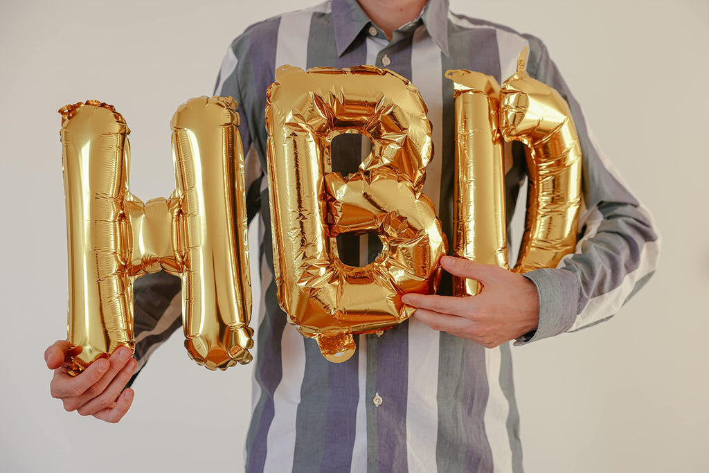 Person holding gold foil letter balloons spelling 'HBD'