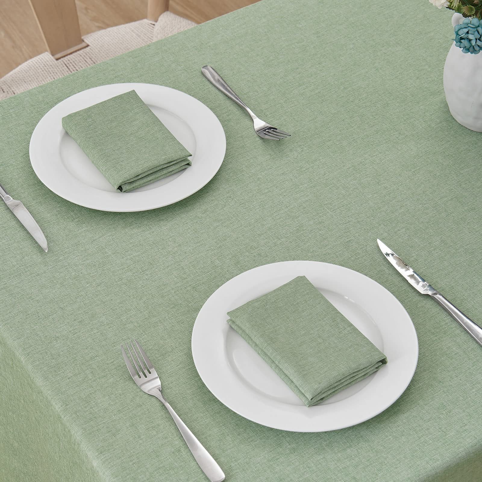 Sage green linen textured tablecloth set with place settings for two, including plates, napkins, forks, and knives.