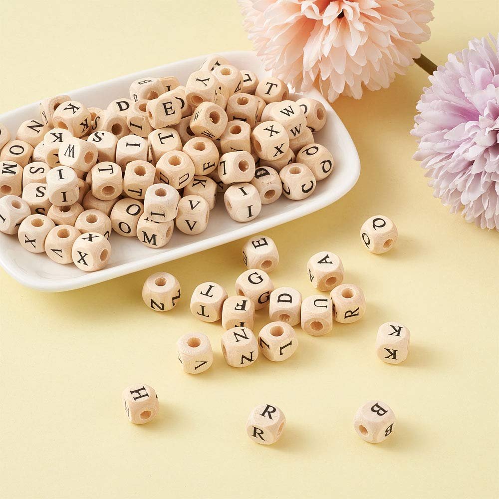 Natural wood cube alphabet beads scattered on a yellow surface with a white dish and decorative flowers.