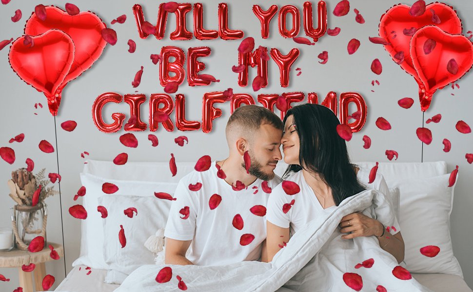 Couple in bed surrounded by red heart balloons and rose petals, with 'WILL YOU BE MY GIRLFRIEND' balloons overhead.