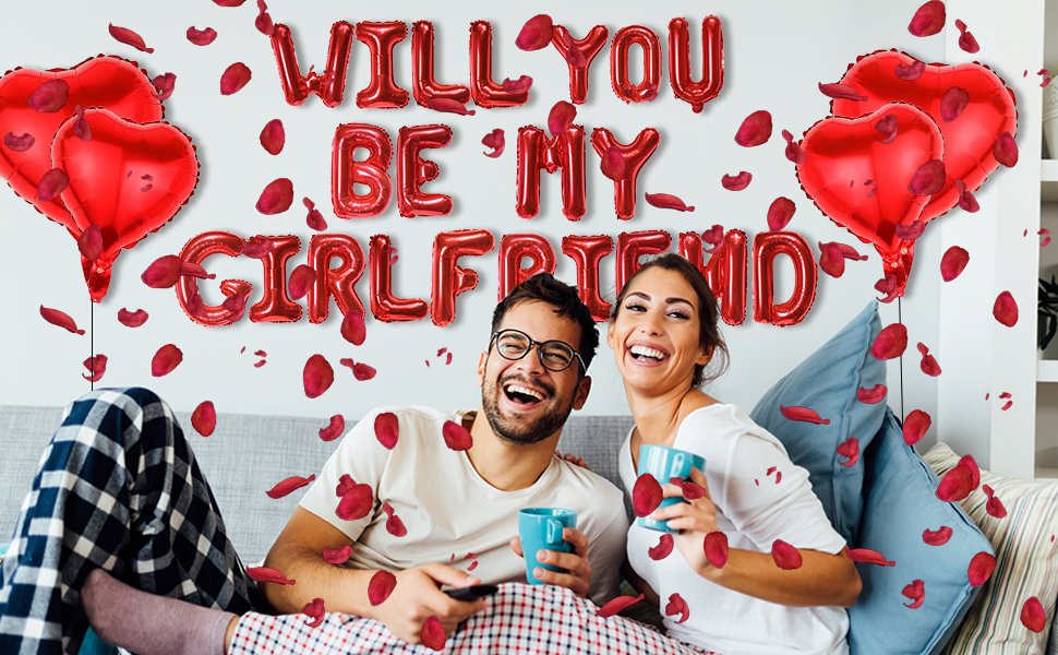 Couple laughing on a couch surrounded by red heart balloons and rose petals, with 'WILL YOU BE MY GIRLFRIEND' balloons overhead.