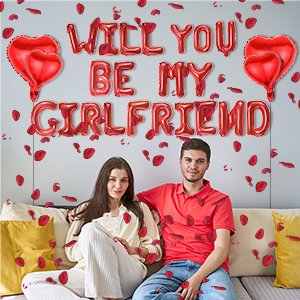 Couple sitting on couch surrounded by red heart balloons and rose petals, with 'WILL YOU BE MY GIRLFRIEND' balloon text above.