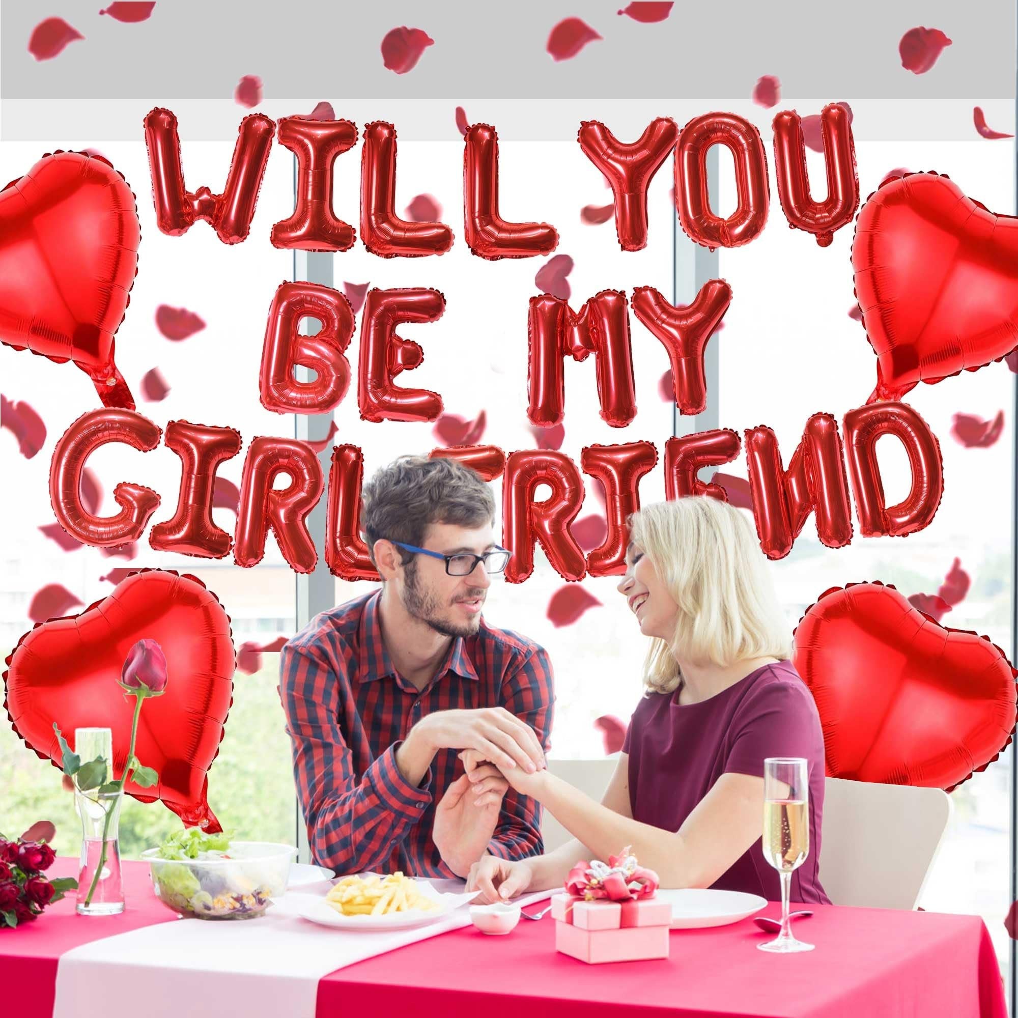 Couple celebrating a proposal with red heart and letter balloons that spell 'Will You Be My Girlfriend'