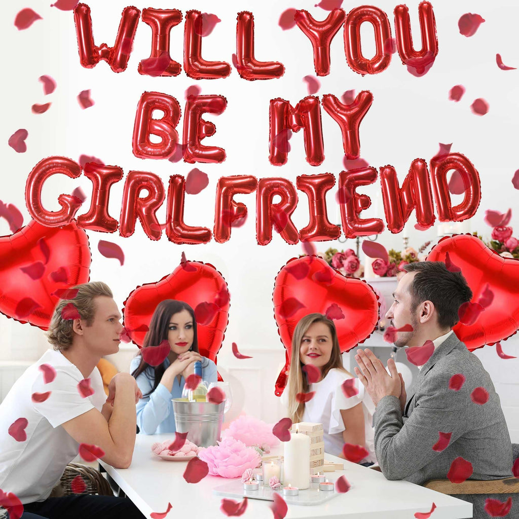 Four people at a table with red heart and letter balloons spelling 'Will You Be My Girlfriend', rose petals scattered.