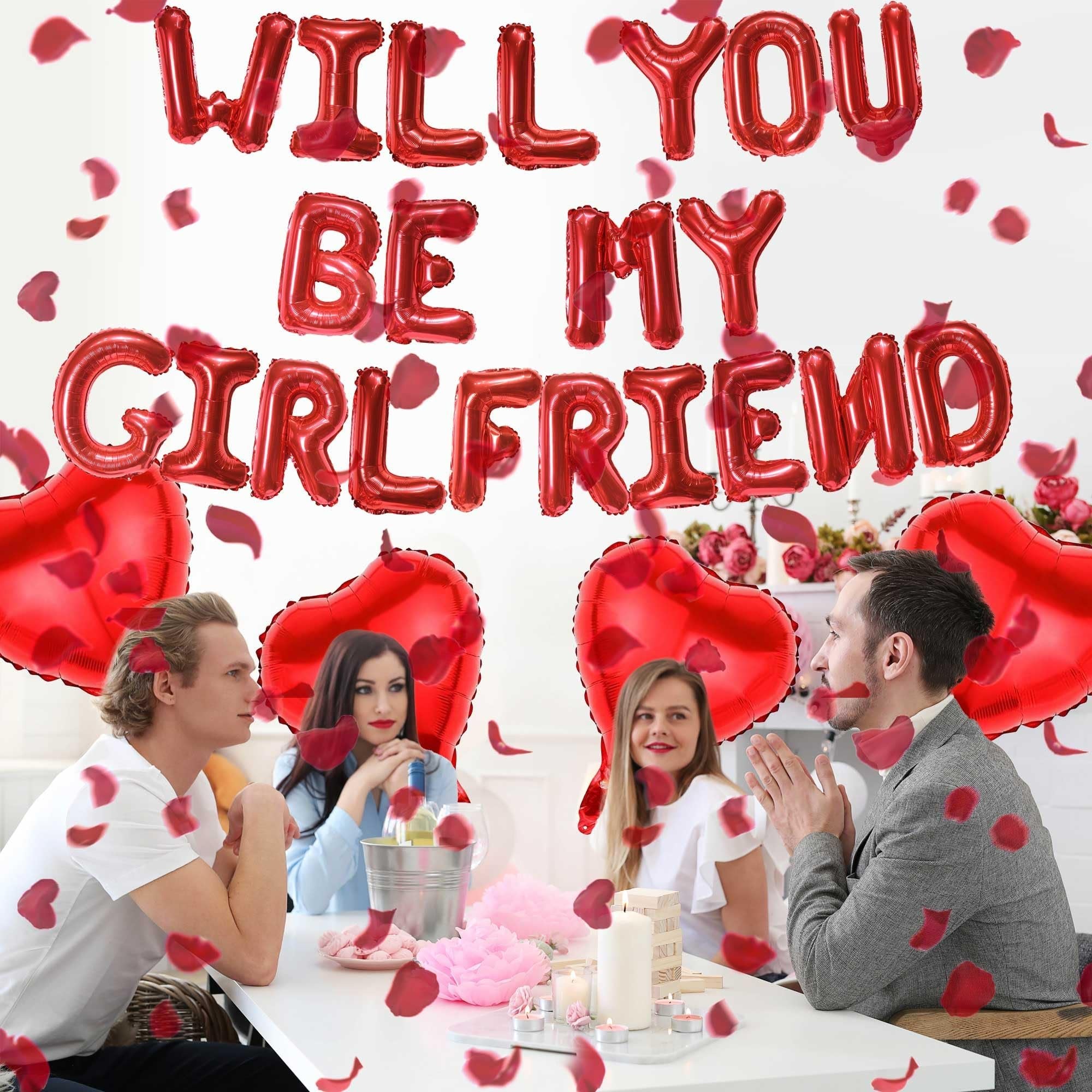 Four people at a table with red heart and letter balloons spelling 'Will You Be My Girlfriend', rose petals scattered.