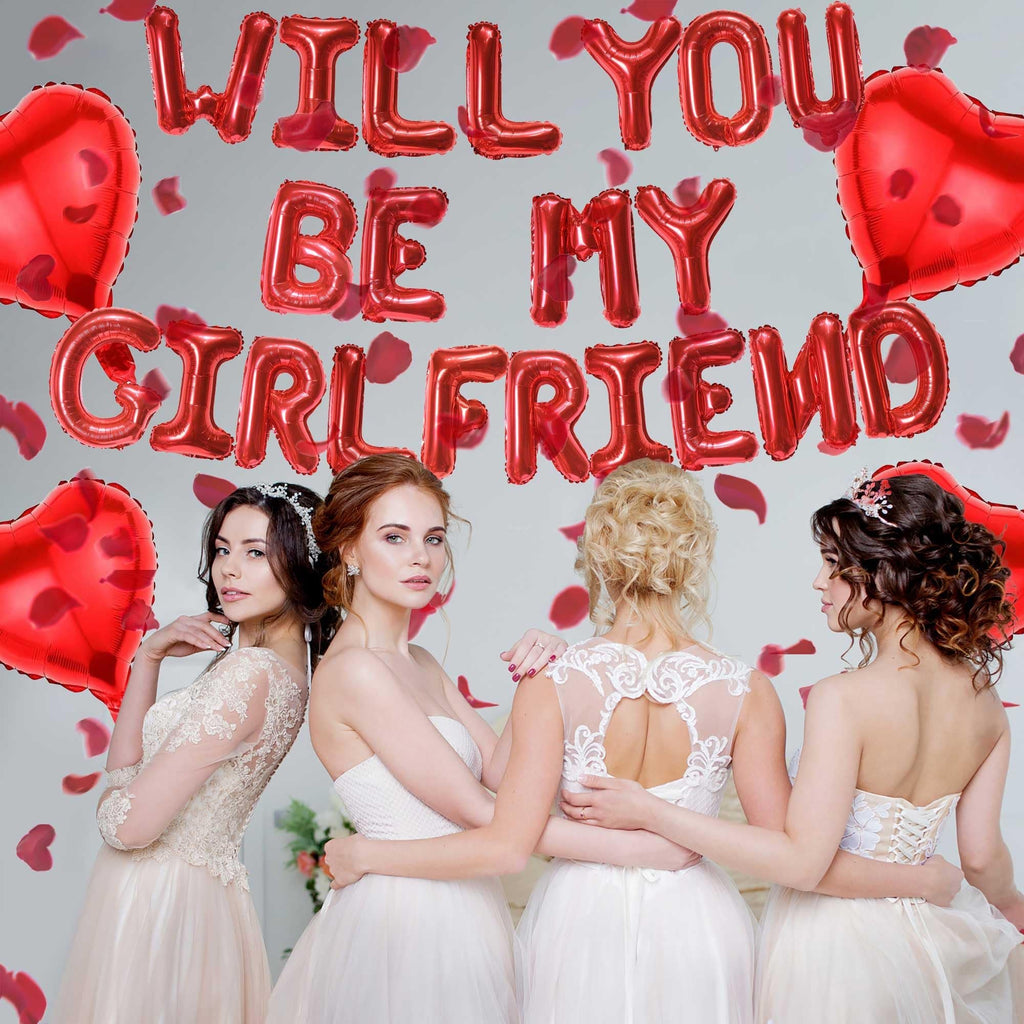 Four women in white dresses posing with red heart and letter balloons that spell 'WILL YOU BE MY GIRLFRIEND'