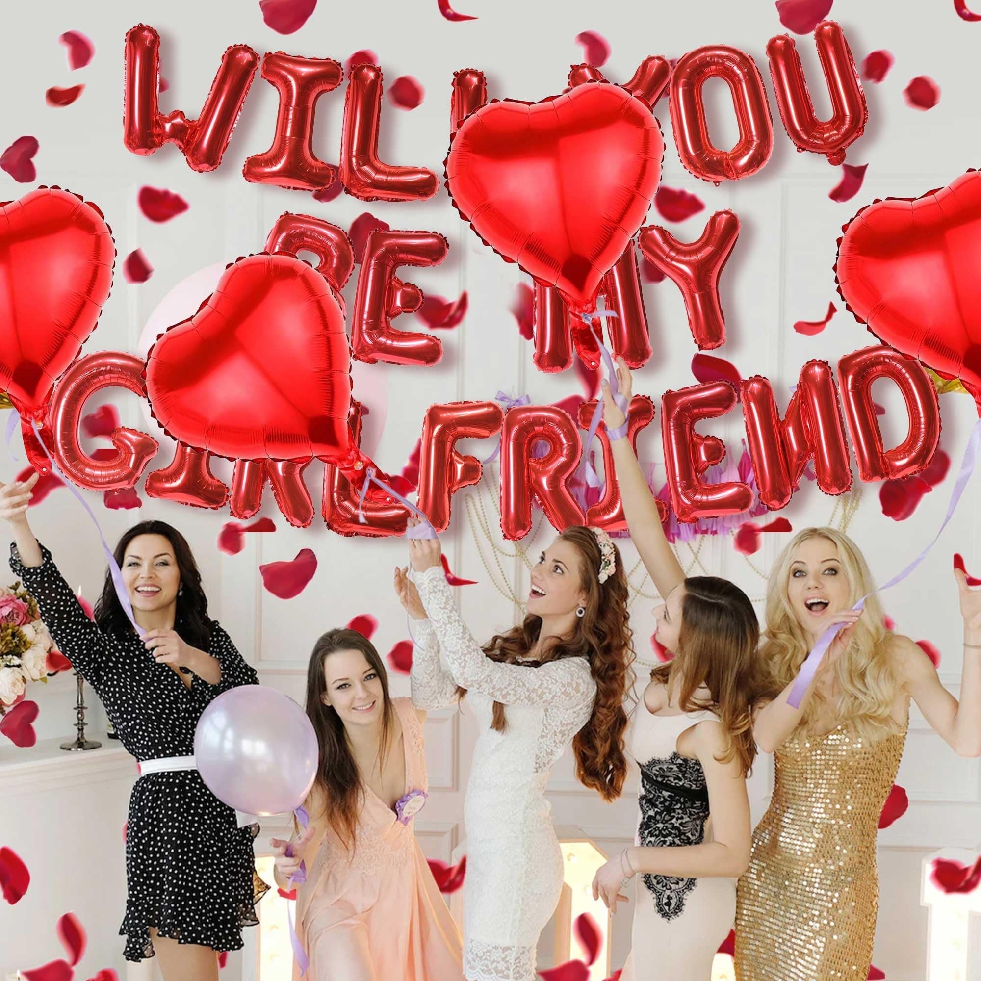 Group of women celebrating with red heart and letter balloons that spell 'Will You Be My Girlfriend' and scattered rose petals.