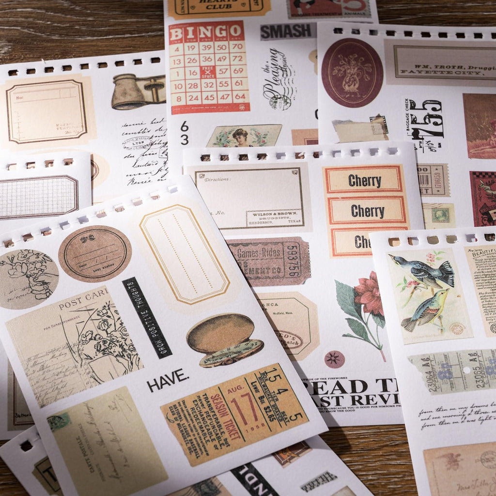 A flat lay of vintage-style scrapbook stickers featuring various designs like bingo cards, tickets, postcards, and floral illustrations on a wooden background.