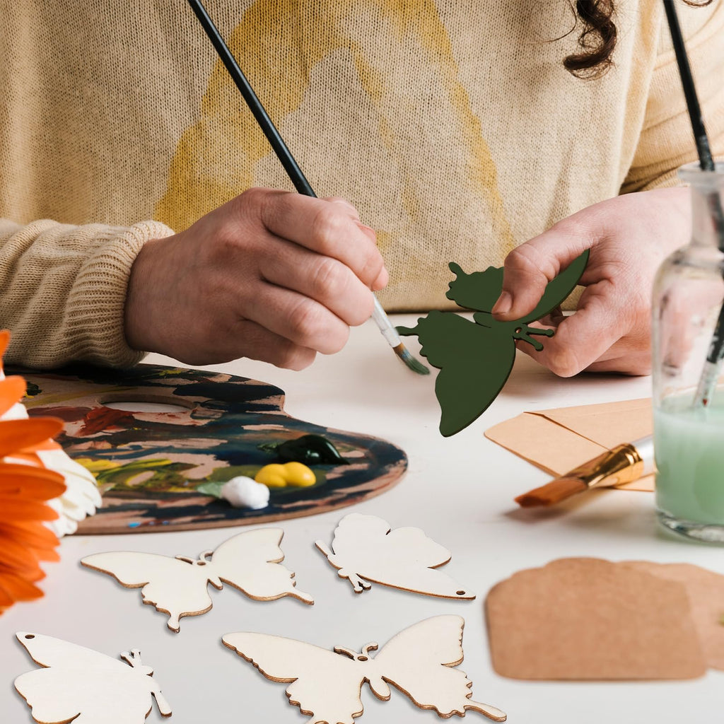 Person painting a dark green wooden butterfly cutout with a brush and palette, surrounded by unfinished wooden butterfly cutouts.
