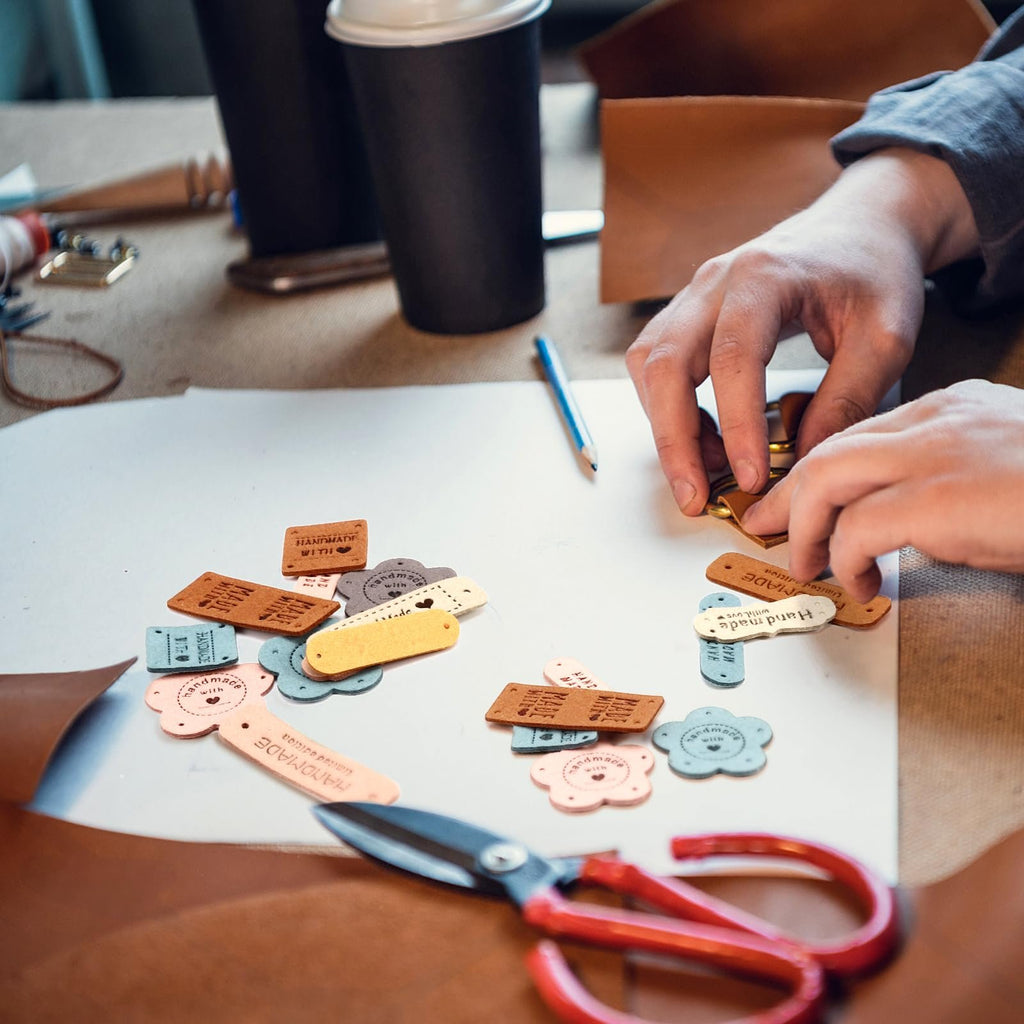Assortment of colorful handmade leather labels with pre-drilled holes scattered on a white surface, with hands sorting them.