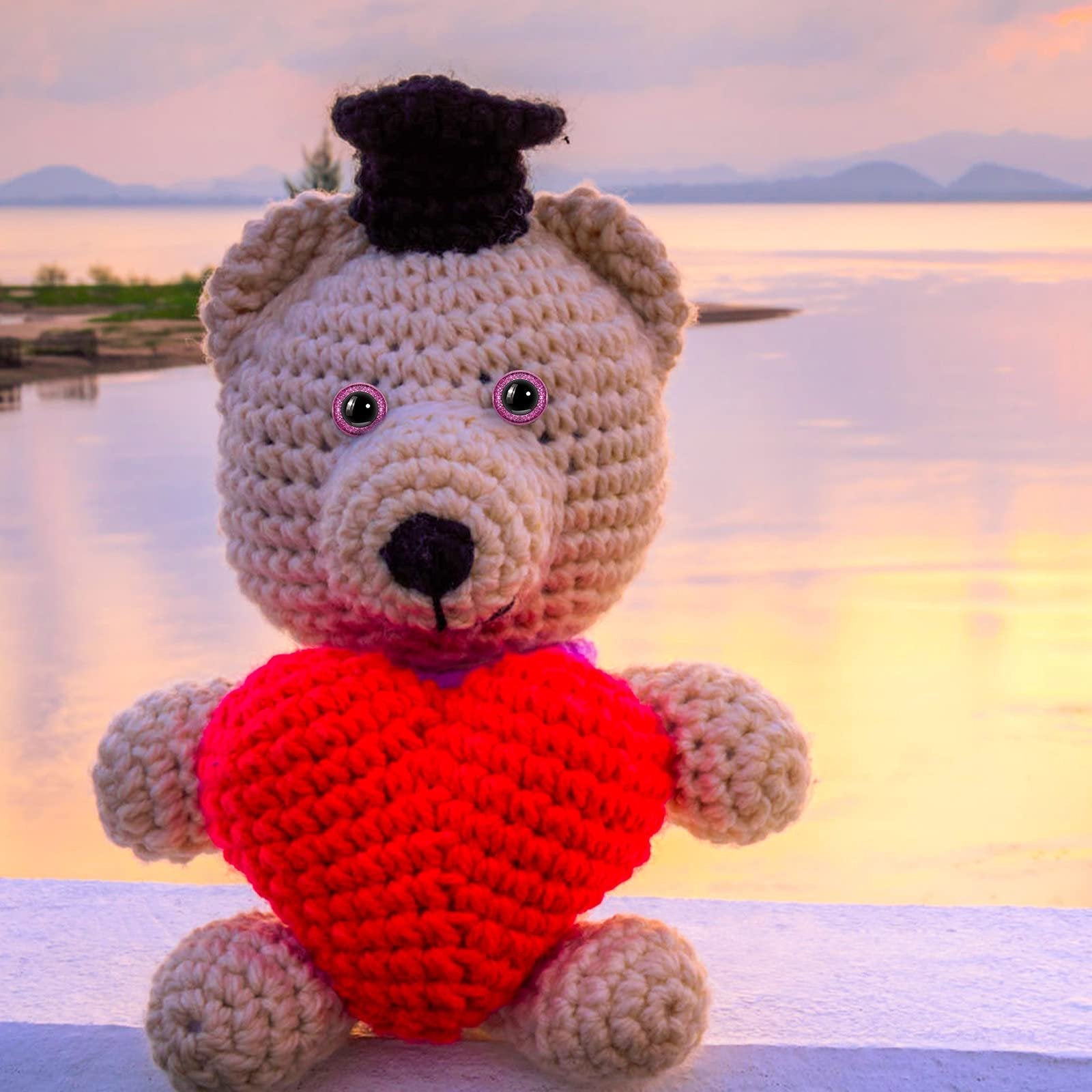 Crocheted teddy bear with red heart and graduation cap against a sunset lake background