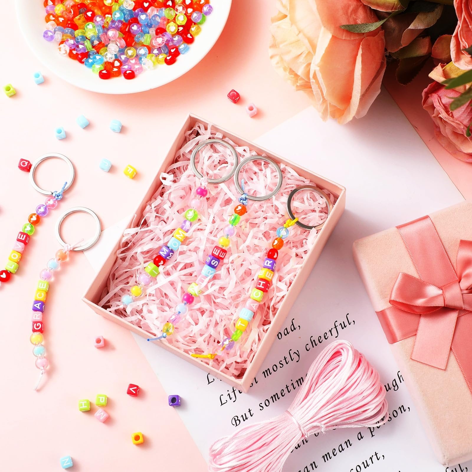 DIY bead keychain kit with letter beads spelling GRACE and LOVE, colorful beads, and metal rings, arranged on a pink background with flowers.