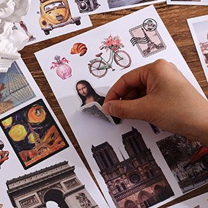 Hand peeling a Mona Lisa sticker from a sheet of vintage travel scrapbook stickers featuring landmarks and icons.